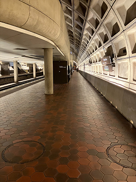 A picture of the pathway further down the platform. To the left there is a side wall, to the right there are train tracks. In the background there is one yellow wet floor cone, as well as the structure for some escalators slightly to the right of center.